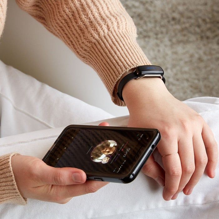 A person holding a smartphone showing the Bond Touch app, wearing a bracelet with a silicone band and a sleek, elongated module piece.