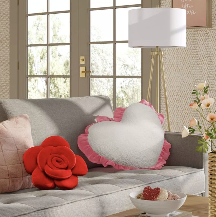 A cozy living room featuring a gray sofa adorned with heart-shaped and rose-patterned pillows, a standing lamp, and a side table with fresh flowers and a small bowl.