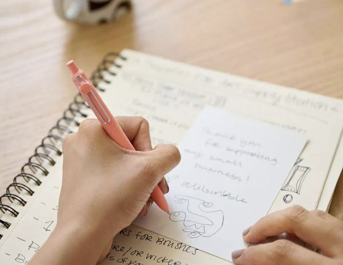 A person writing a note with a pen on paper, using a notebook underneath. A nearby cup is partially visible.