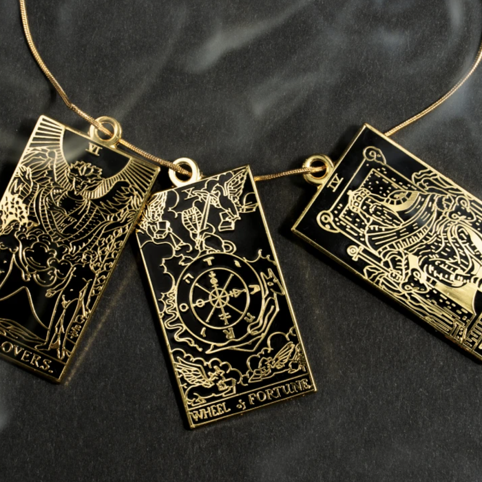 A close-up of three potential tarot charms, each on a chain: The Lovers, The Wheel of Fortune, and The Emperor.