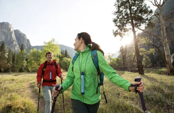Two hikers trekking in the mountains, smiling and carrying trekking poles, both equipped with Lifestraws.