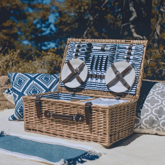 An open wicker picnic basket, neatly displaying plates and utensils, surrounded by decorative pillows in a laid-back outdoor setting