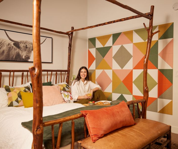 A person beams while sitting on a rustic wooden bed in a room adorned with geometric wall art and a striking cow print. The ambiance is both stylish and contemporary.