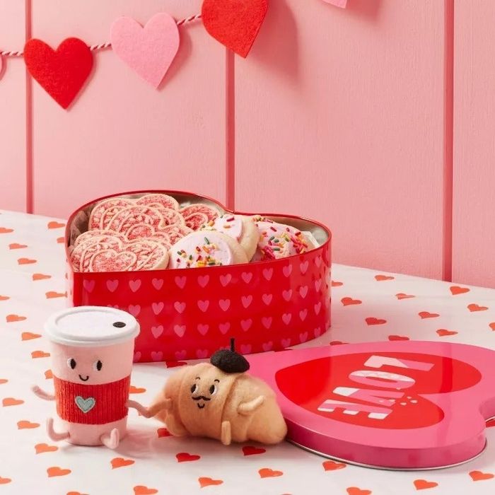 A heart-shaped tin box filled with cookies, alongside plush coffee cup and croissant, set on a beautifully decorated table with a Valentine's Day theme.