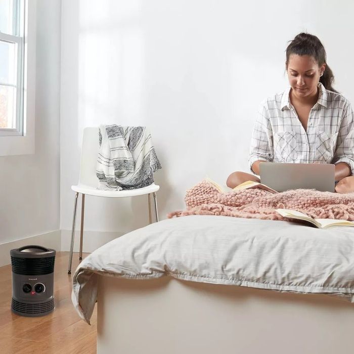 A person sits on a bed, wrapped in a blanket and using a laptop, while a space heater sits on the floor beside them.