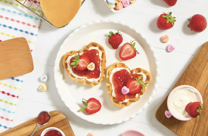 Heart-shaped waffles topped with whipped cream and strawberries on a white plate, surrounded by fresh strawberries and candy hearts on a wooden table