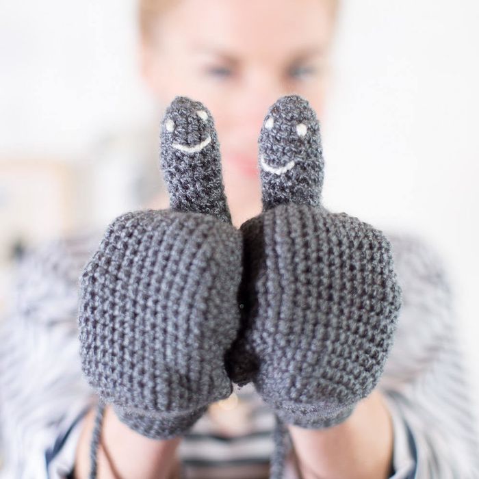 Model wearing grey crocheted mittens with white smiley faces on the thumbs