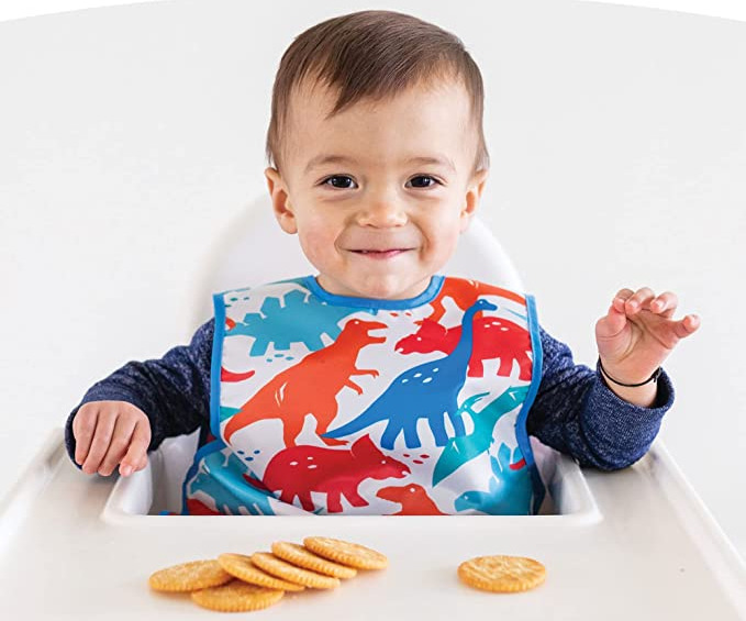 A young child sitting in a highchair, sporting a dinosaur-themed apron bib, munching on crackers.
