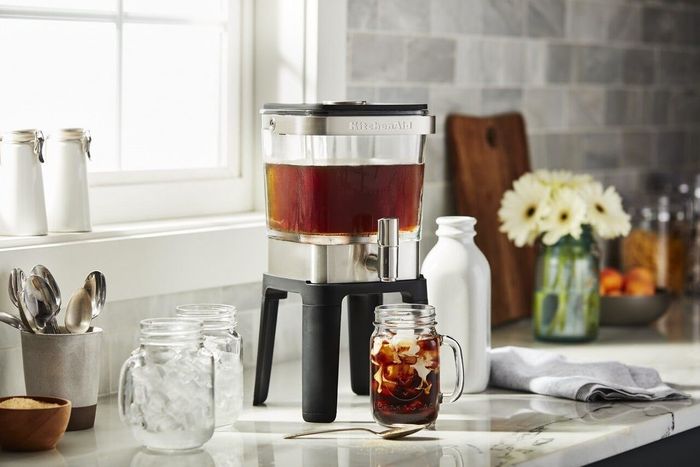 Glass and metal cold brew coffee maker sitting on a kitchen counter, accompanied by iced coffee in a mason jar and nearby kitchen essentials
