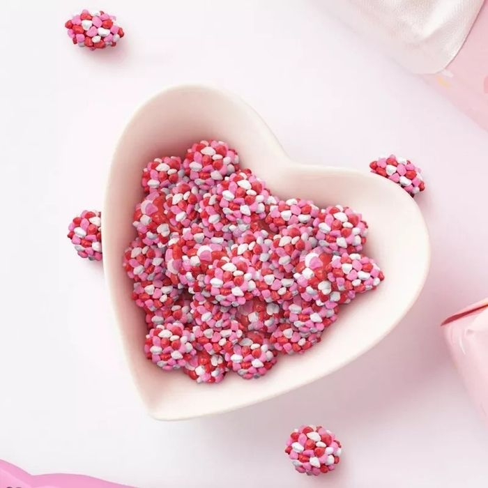 A heart-shaped bowl filled with small candy clusters, adorned with colorful sprinkles. Additional candies are scattered around the bowl, completing the sweet display.