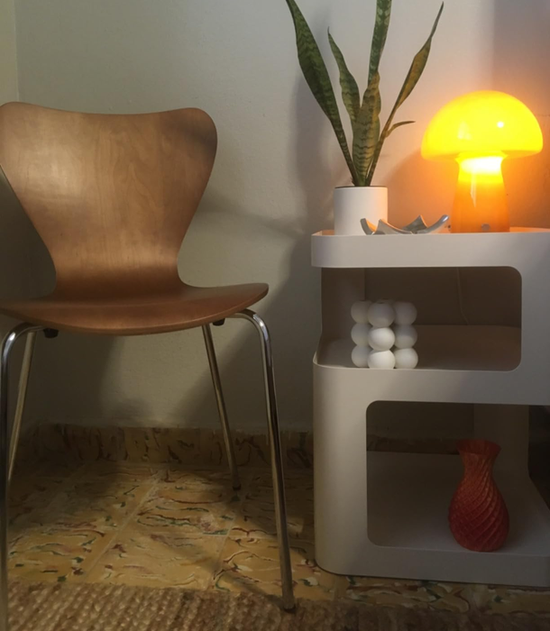 A minimalist room featuring a wooden chair, a small white table with a potted snake plant, a yellow lamp, and a red vase placed on the bottom shelf.