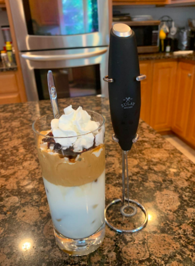 A reviewer using a black milk frother to prepare Dalgona coffee in their kitchen