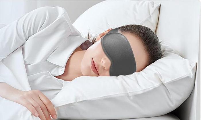 A person wearing a sleep mask, resting on a bed with white sheets, featured in a shopping article on sleep accessories.