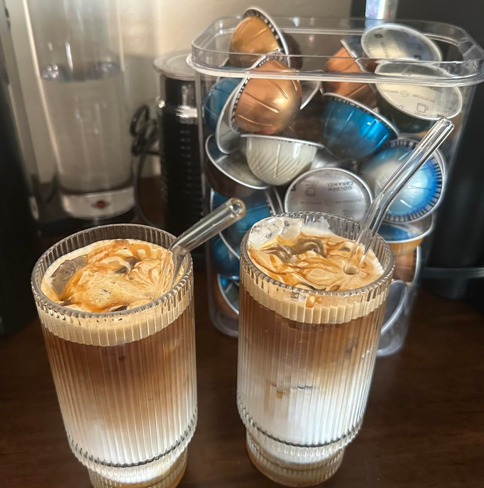 Two iced coffee drinks with glass straws next to a container packed with a variety of coffee pods.
