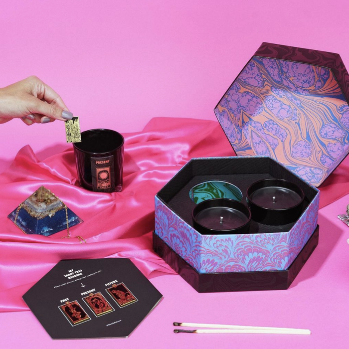 A beautifully designed box with three tarot-themed candles inside. Alongside the candles, there are a crystal, charms, and a hand pulling a charm from one of the melted candles.