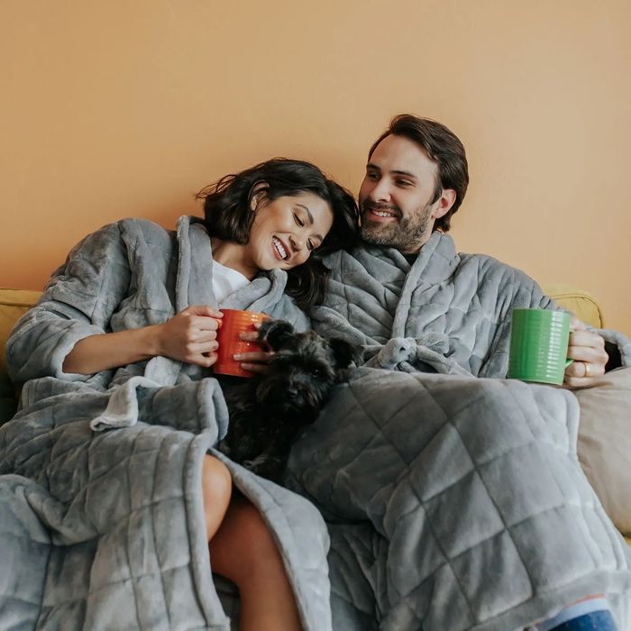 A couple, both wearing plush robes, smiles while holding mugs on a couch with a small dog.