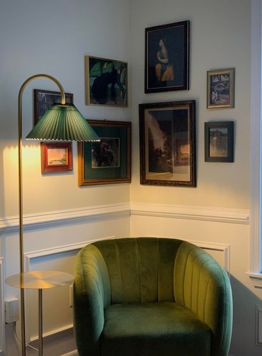 A comfortable reading nook with a green velvet chair, a gold floor lamp, and a gallery wall filled with various framed art pieces.