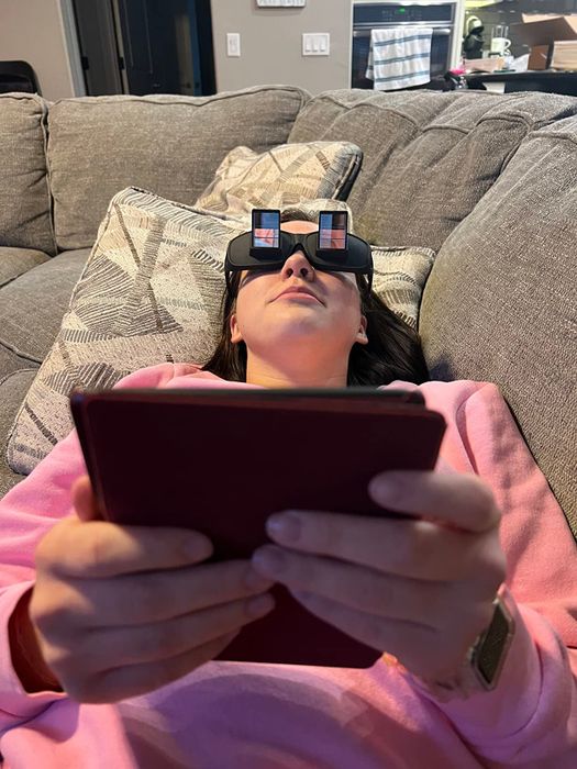 A person lying on a couch, wearing prism glasses while holding a tablet.
