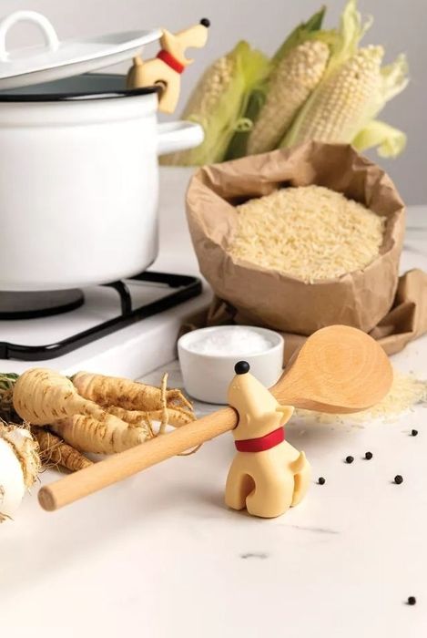 A wooden dog-shaped spoon holder keeping a wooden spoon in place on the kitchen counter, surrounded by rice, corn, parsnips, and a cooking pot.