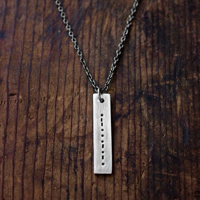Metal plate necklace on a chain in a long rectangular shape, featuring morse code dots and dashes pressed into the plate.