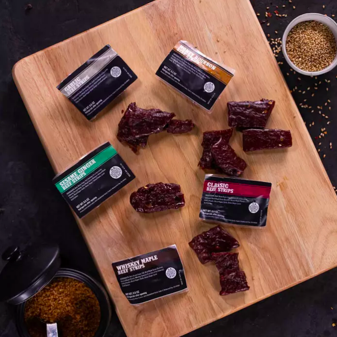Various pieces of jerky arranged on and around their packaging on a cutting board.