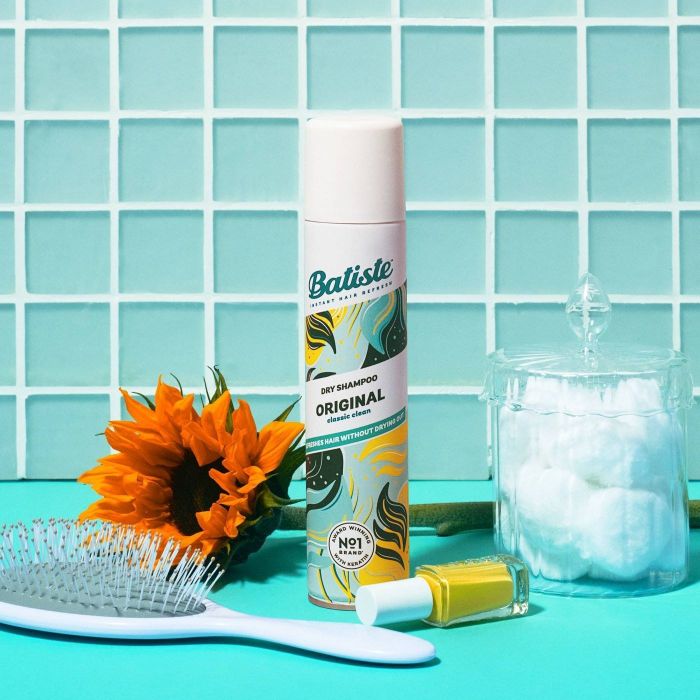 A bottle of blue, green, and yellow dry shampoo placed on a blue table alongside a blue hairbrush, a sunflower, yellow nail polish, and a jar of cotton puffs