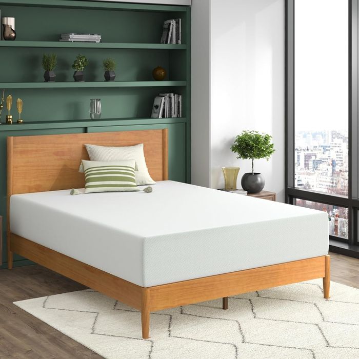A modern bedroom featuring a wooden bed frame, a white mattress, a striped pillow, and a shelf adorned with plants and trophies. A large window offers a breathtaking view of the city in the background.