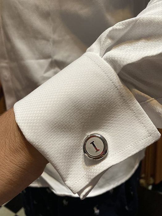 The round silver cufflinks featuring the letter I initial, attached to a shirt cuff.