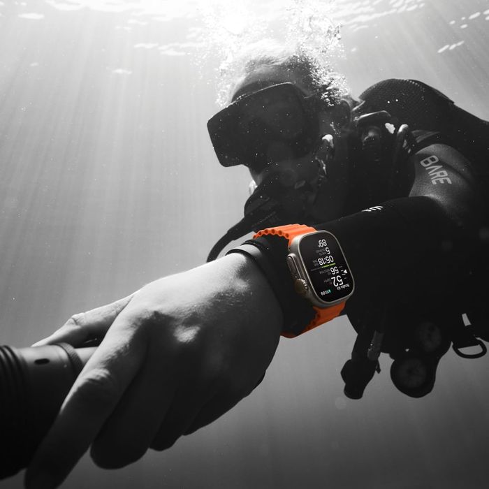 A person in scuba gear submerged underwater, wearing an Apple Watch Ultra with an orange strap. The watch face displays diving metrics.