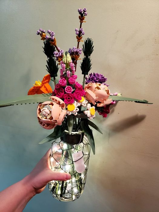 A hand holding a vase with Lego flowers inside, adorned with fairy lights.