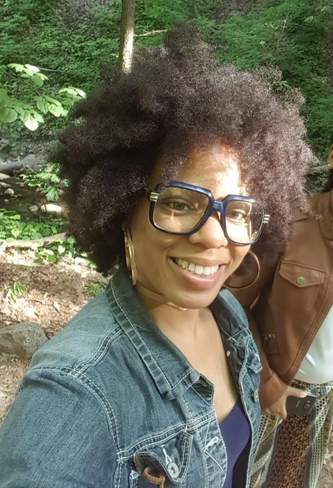 A reviewer wearing a denim jacket with brunette, kinky hair standing outdoors.
