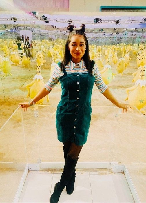 A reviewer dressed in a dark green pinafore dress layered over a collared shirt