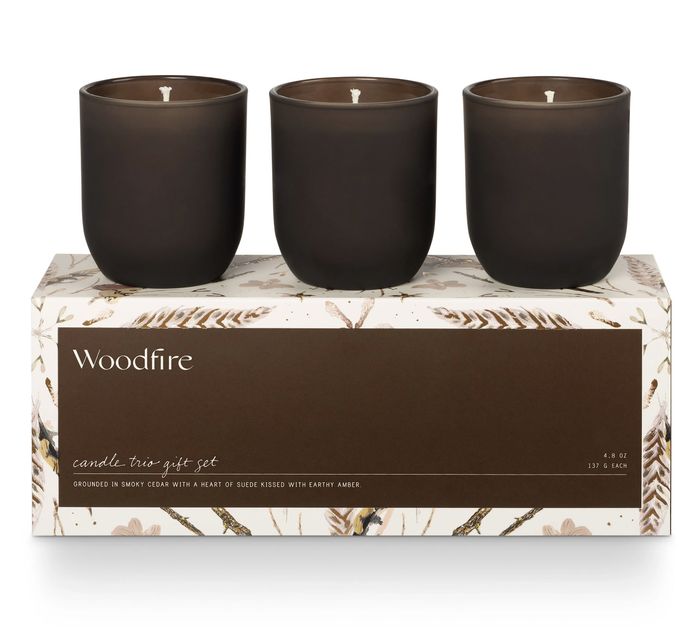 Woodfire candle trio gift set in brown containers, resting on a decorative box, emitting scents of cedar, suede, and amber