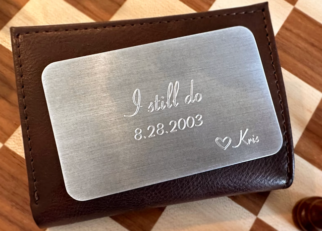 A slim aluminum card, shaped like a credit card, etched with the words 'I still do,' the wedding date, a heart, and the name Kris at the bottom.