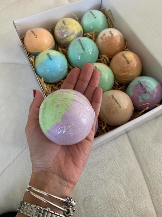 A reviewer's hand with a large bath bomb, surrounded by a box filled with colorful bath bombs.