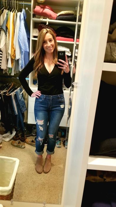 A person takes a mirror selfie in a closet, wearing a black top, ripped jeans, and ankle boots, showing off a casual outfit option.