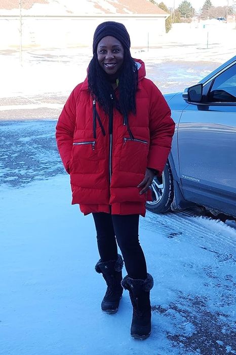 A person stands on a snowy driveway, dressed in a winter coat, hat, and boots, with an SUV parked nearby.
