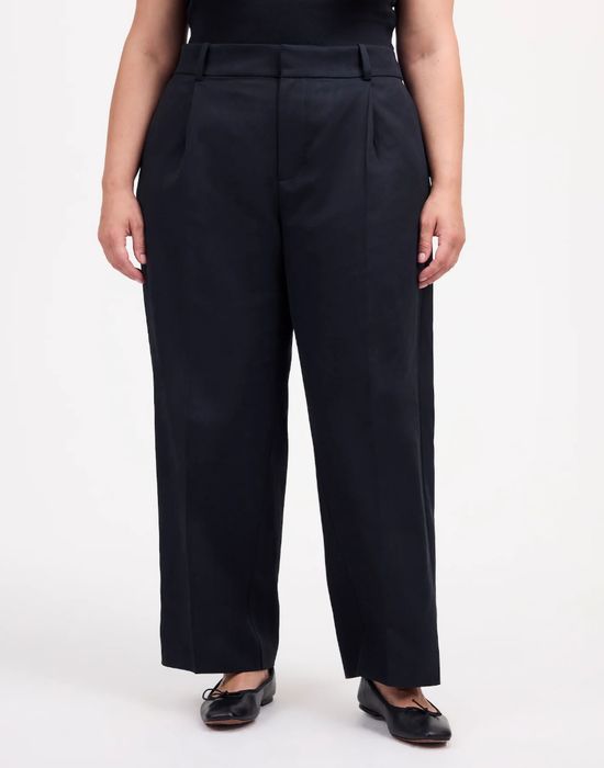 A person wearing relaxed black trousers and black shoes, posing in front of a neutral backdrop.