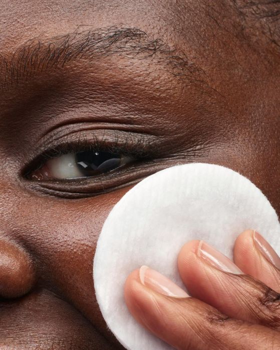 A person smiling while holding a cotton pad to their face, showing the application of skincare.
