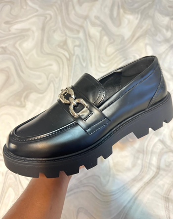 A chunky black loafer with a thick sole and silver chain detail, held in someone's hand for consideration