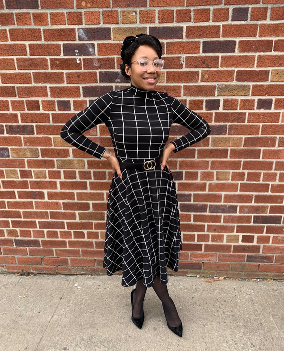 A reviewer wearing a black and white high-neck plaid dress with long sleeves, paired with a black and gold belt, black tights, and black pointed-toe heels, standing in front of a brick wall.