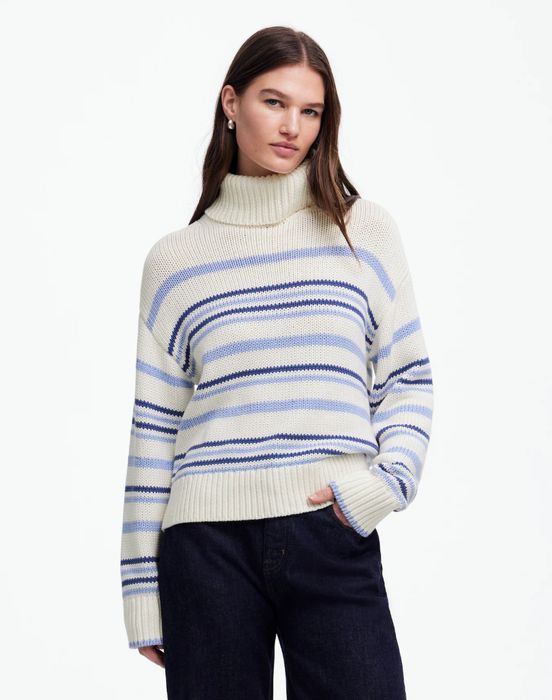A person dressed in a striped turtleneck sweater and dark pants, casually standing with one hand in their pocket