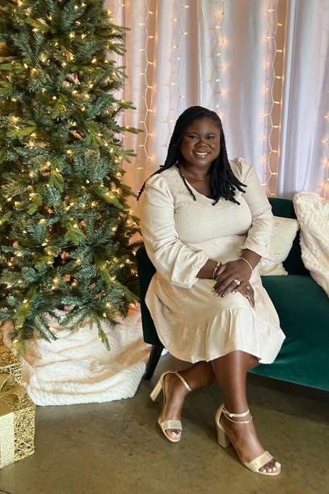 A reviewer wearing a white dress is seated on a green couch next to a beautifully decorated tree and wrapped presents.