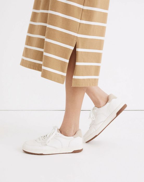 A person is wearing a striped skirt and white sneakers while standing on a plain white floor, showcasing a simple and stylish look.