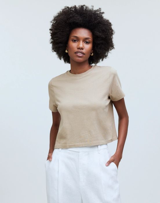A person with curly hair, wearing a beige T-shirt and white pants, confidently posing with their hands in their pockets