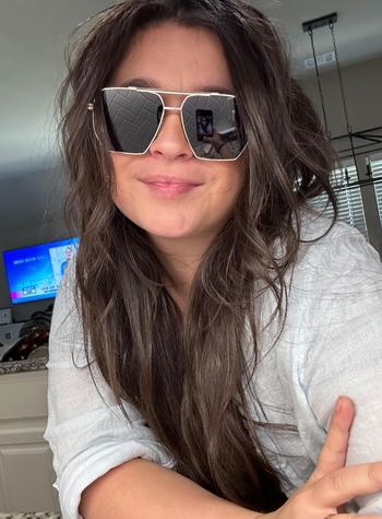 A person with long hair wearing oversized sunglasses and a light blouse indoors, with a TV visible in the background