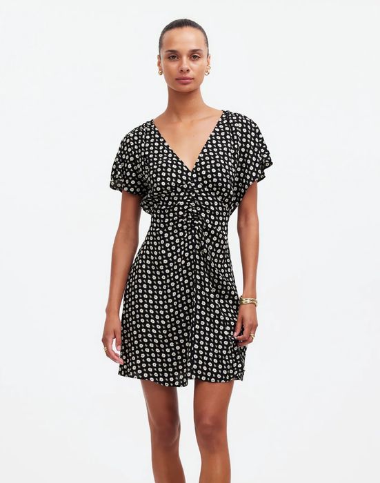 A person is styled in a patterned short-sleeve wrap dress, accessorized with hoop earrings and bracelets, standing against a neutral background.