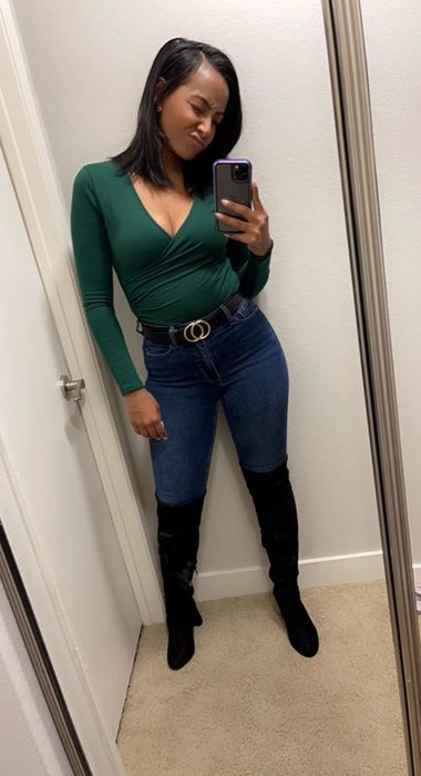 A reviewer takes a mirror selfie, wearing a green wrap top, blue jeans, a black belt, and knee-high black boots.