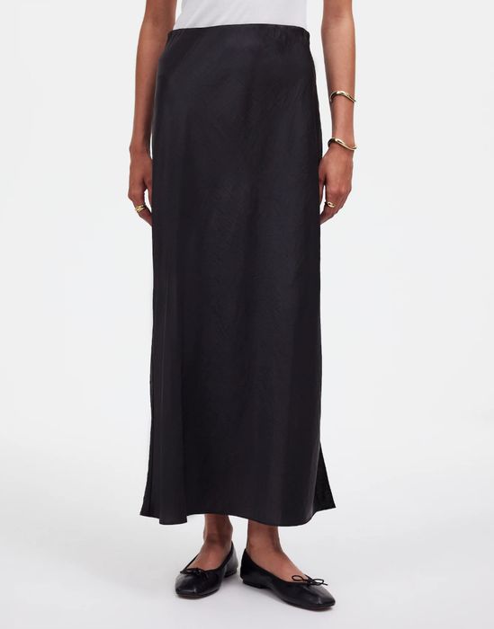 Person wearing a long, chic black skirt paired with a white top, accessorized with simple bracelets and black flats.