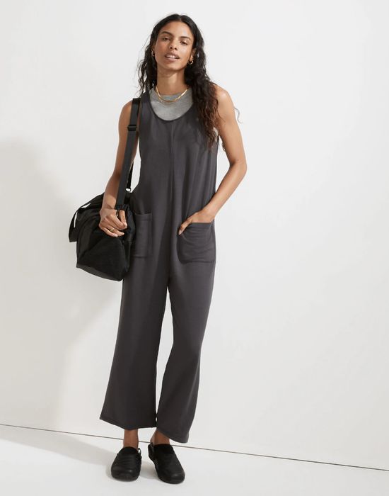 A person in a casual jumpsuit with pockets, carrying a shoulder bag and wearing a bold statement necklace, stands against a minimalist background.